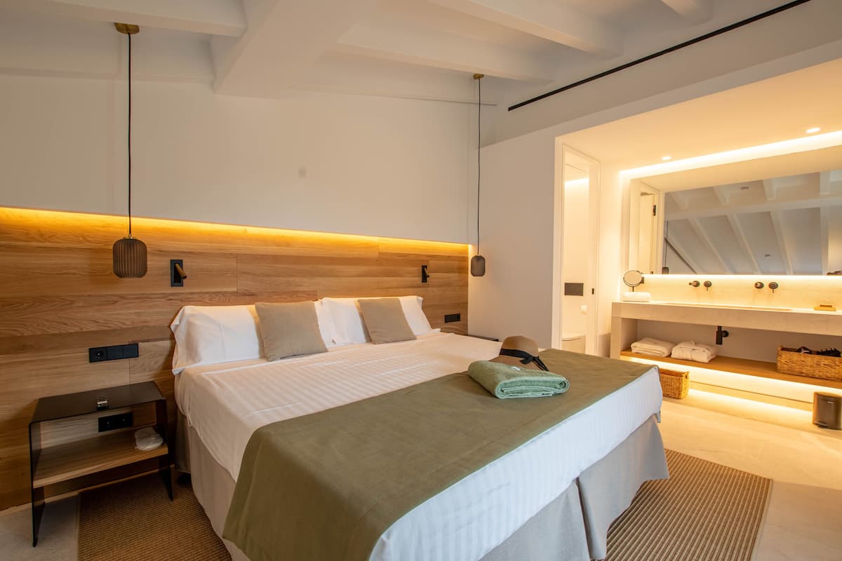 Un dormitorio moderno con cabecera de madera, una cama grande con ropa blanca, una mesita de noche y un baño con un espejo grande y un perchero para toallas.