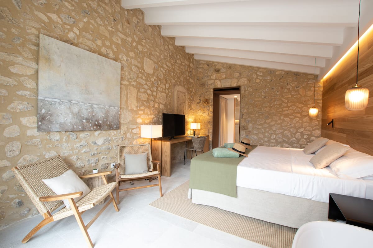 Una habitación con pared de piedra, una cama, un escritorio con televisor, una silla de mimbre y un cuadro grande.