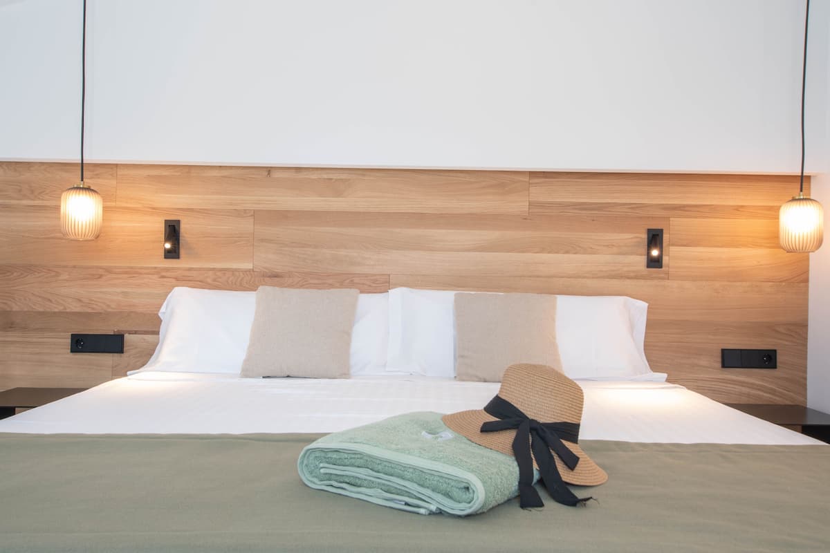 Una cama bien hecha con una toalla doblada y un sombrero, cabecero de madera y dos lámparas colgantes.