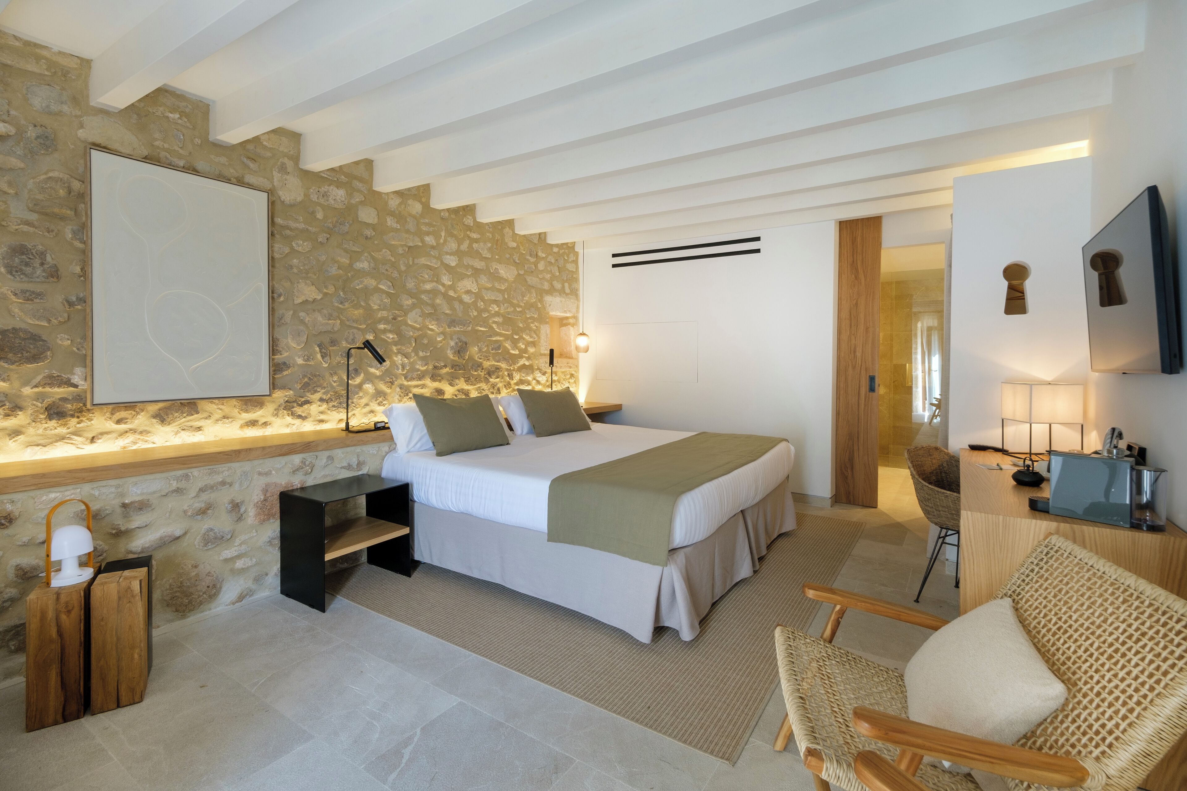 Un dormitorio con pared de piedra, una cama con colcha color beige, una mesita de noche de madera, una silla de mimbre y un escritorio con lámpara.