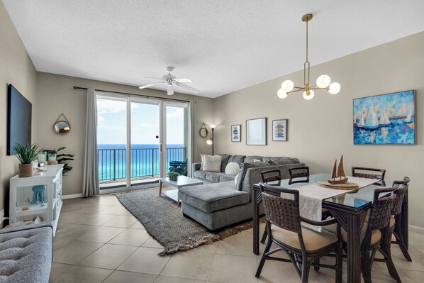 A spacious living room with a grey sofa, a dining table with wicker chairs, a ceiling fan, and a large window overlooking the ocean.