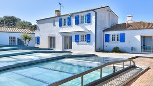 Una piscina con cerramiento de vidrio, un edificio blanco con contraventanas azules y un techo de tejas.