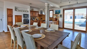Un comedor espacioso con una gran mesa de madera dispuesta para seis personas, rodeada de sillas de mimbre. Se aprecia una cocina con armarios de madera y un microondas moderno al fondo.