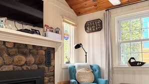 A cozy living room with a stone fireplace, a blue armchair, a yellow bean bag, a wooden table, and a large window with a view.
