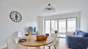 A modern living room with a wooden dining table, white chairs, a blue sofa, and a large window. - Ecapade for 4 in Pléneuf-Val-André (Pléneuf-Val-André)