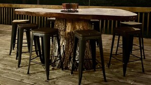 A round wooden table with a central bowl, surrounded by four stools, set on a wooden deck with string lights overhead.