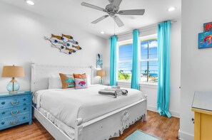 Una camera da letto con un letto bianco, una ventola a soffitto, una finestra con tende, un comodino con una lampada e un quadro appeso al muro.