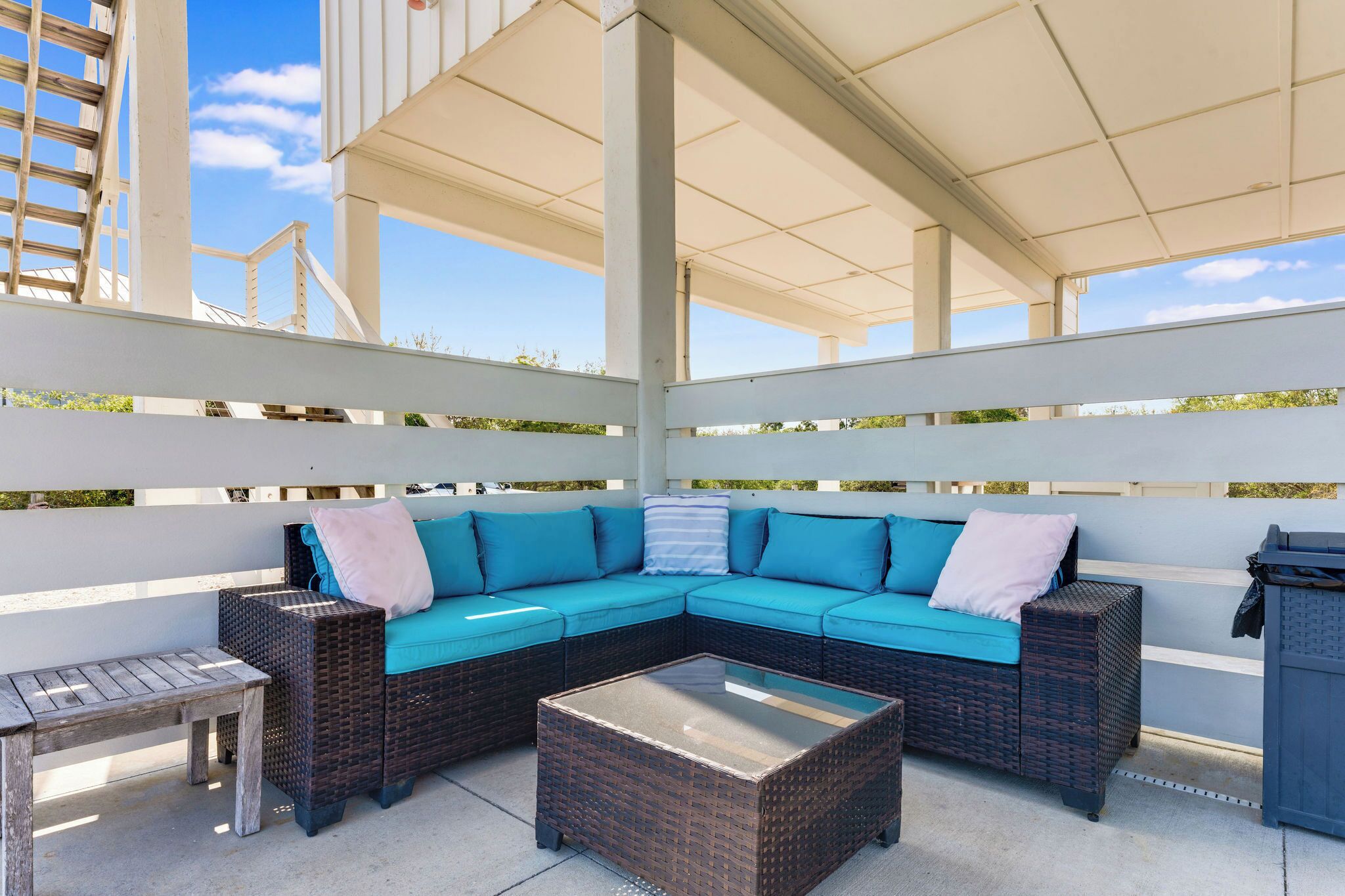 A covered outdoor seating area with a sectional sofa, a small table, and a trash bin.