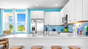 Une cuisine moderne avec des meubles blancs, un réfrigérateur en acier inoxydable, des tabourets en bois et une grande fenêtre donnant sur un paysage verdoyant.