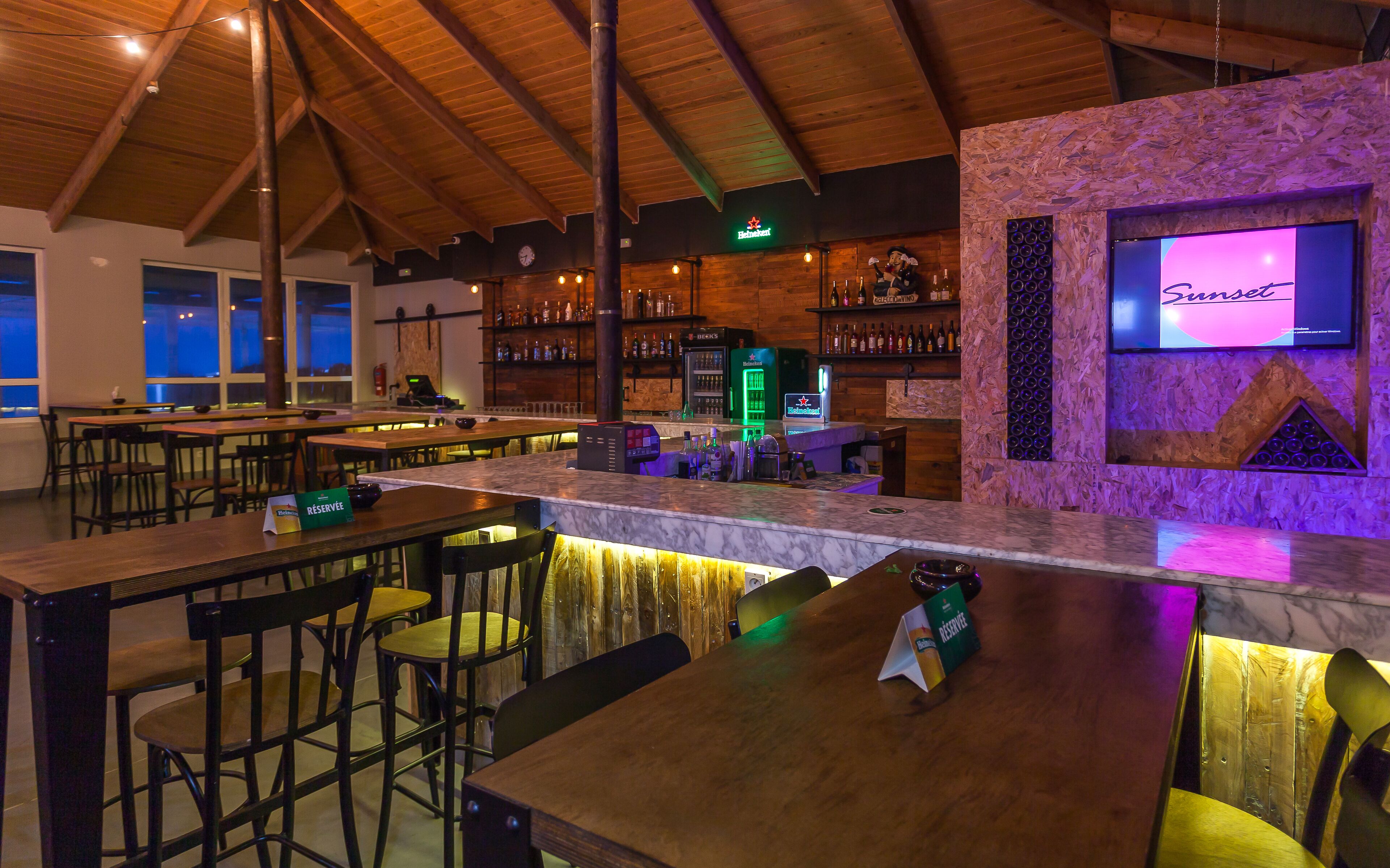 Zona bar con tavoli e sedie in legno, un bancone e scaffali pieni di bottiglie.