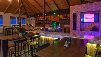 Un área de bar con mesas y sillas de madera, una barra y estantes con botellas.