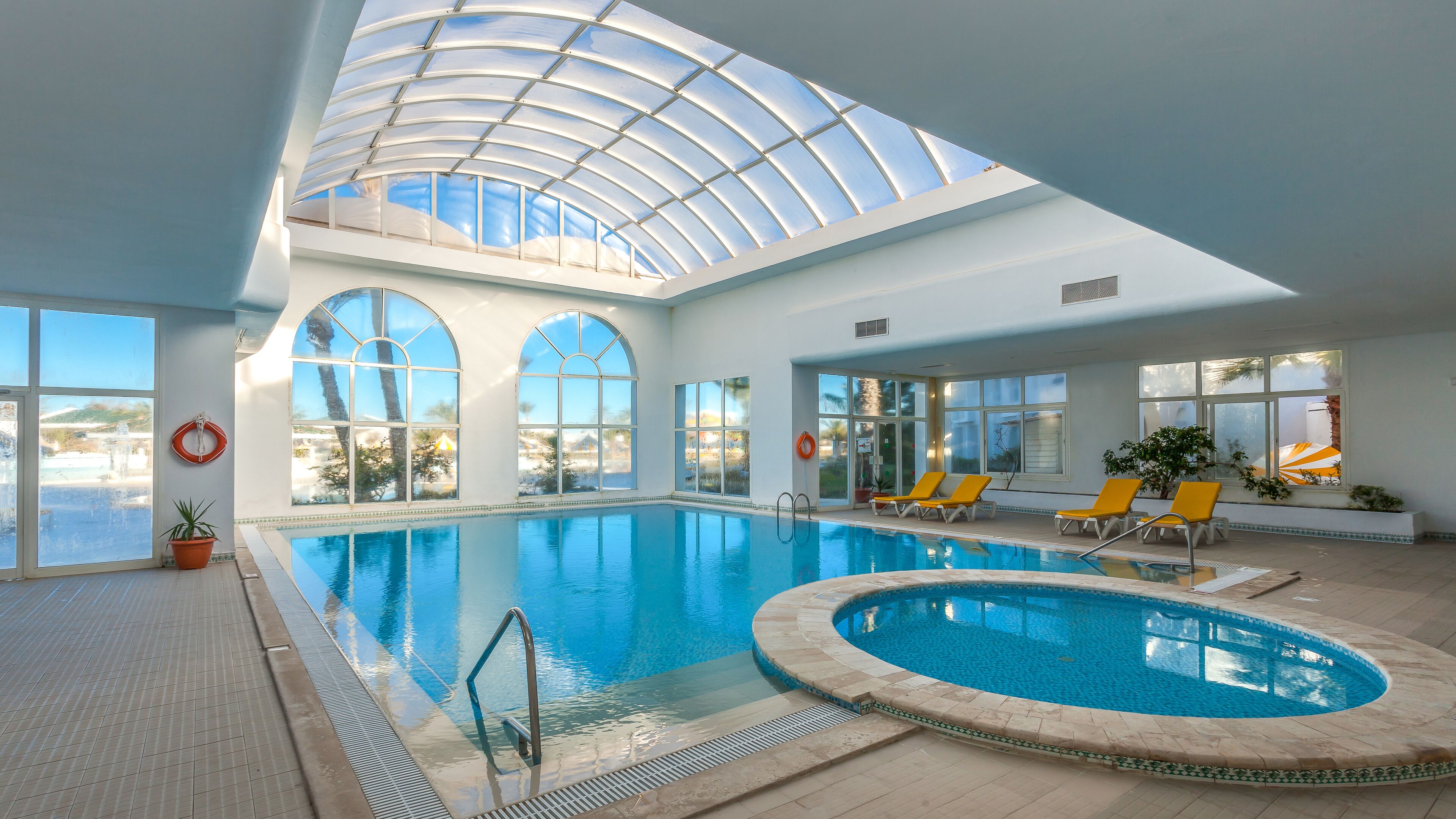 Une piscine intérieure dotée d’un toit en verre transparent, de nombreuses fenêtres et de chaises longues.