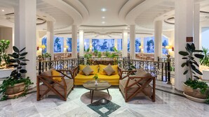 A spacious lobby with a yellow sofa and armchairs, a marble floor, and large windows with a view of palm trees.