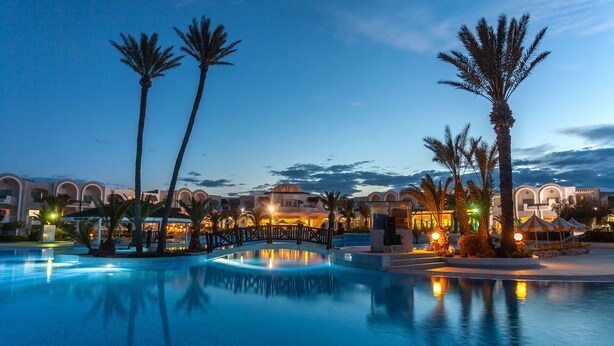 A resort pool area with palm trees, a bridge, and buildings with arched windows.