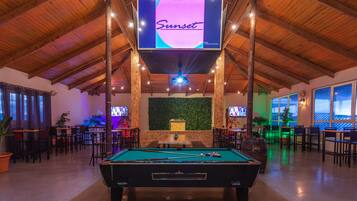 Een pooltafel in een ruimte met houten balken, een televisiescherm waarop 'Sunset' staat en meerdere tafels en stoelen.