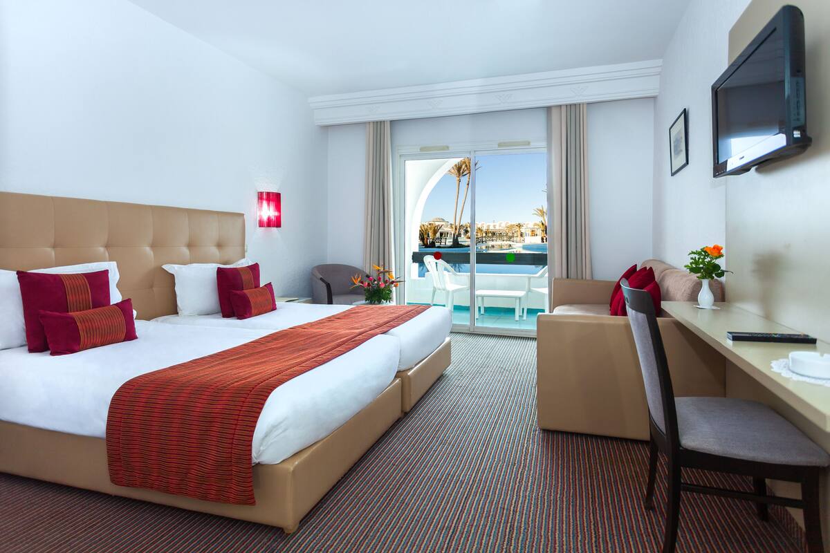 Ein Hotelzimmer mit einem großen Bett, einem Schreibtisch, einem Stuhl, einem Fernseher und Blick auf einen Pool und Palmen.