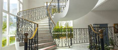 Una imponente escalera con barandillas negras ricamente decoradas y detalles en rojo, que conduce a un segundo piso en un interior amplio, bien iluminado, con ventanas grandes y plantas en macetas.