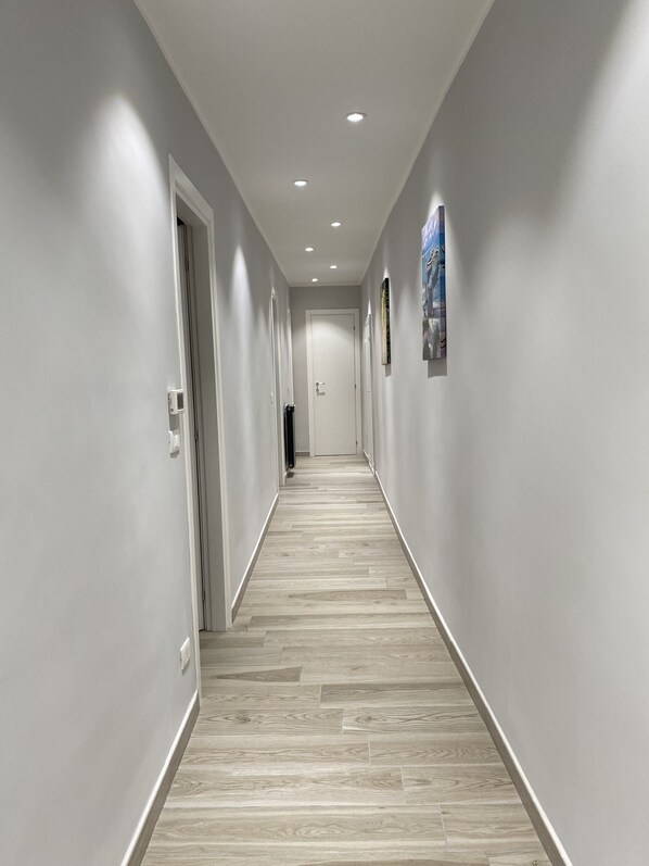 A long, narrow hallway with light-colored wooden flooring and white walls. There are recessed ceiling lights and a door at the end of the hallway.