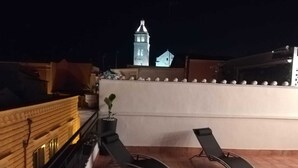 Une terrasse sur le toit avec deux chaises longues, un pot de fleurs et une vue sur un clocher illuminé la nuit.