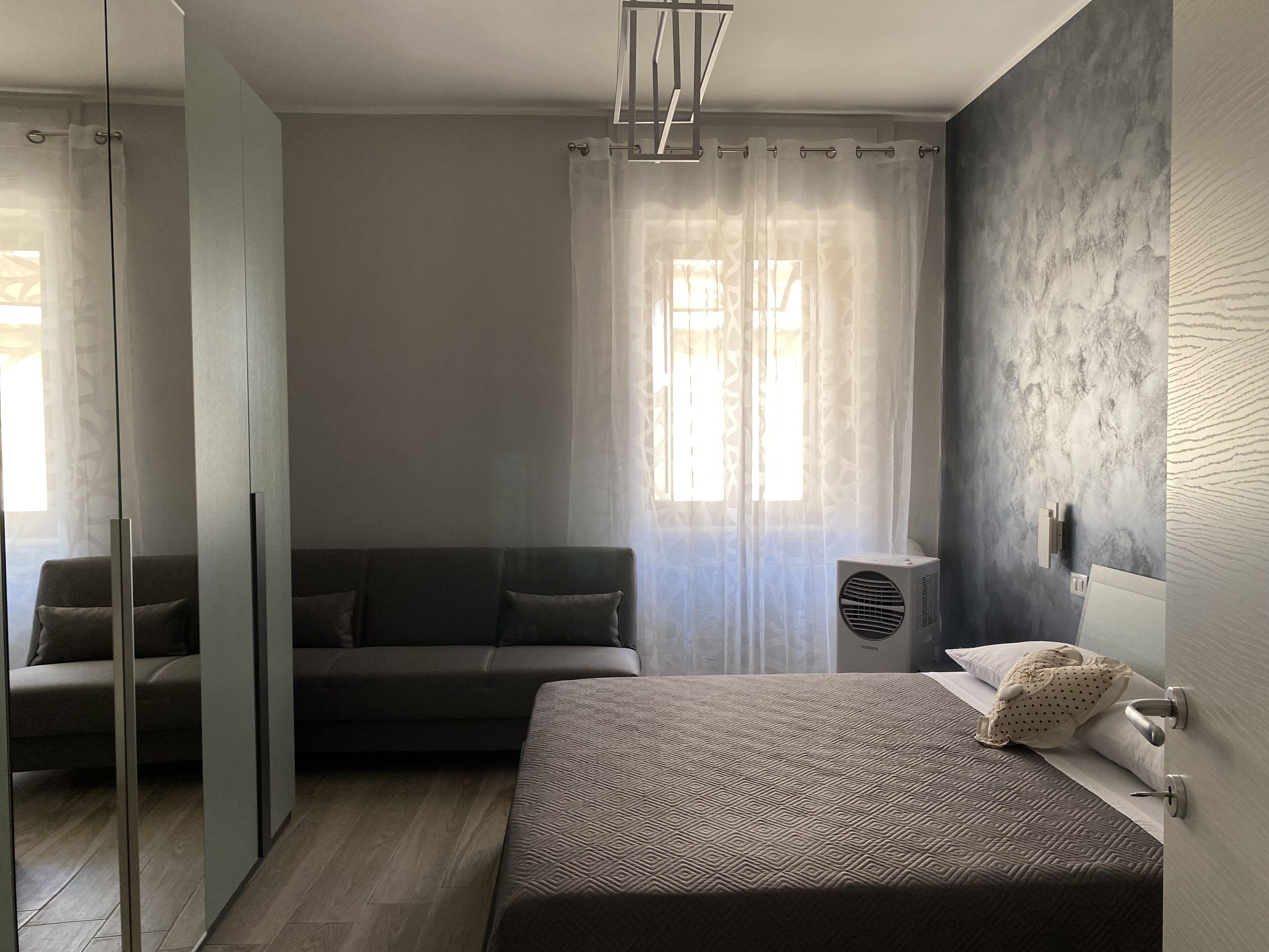 A modern bedroom with a bed, a sofa, a glass partition, and a window with sheer curtains.