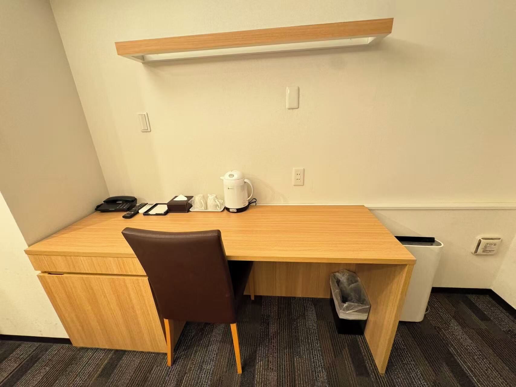 Ein kleines Hotelzimmer mit einem Holztisch, einem Stuhl, einer Kaffeemaschine, einem Telefon und einem Mülleimer.