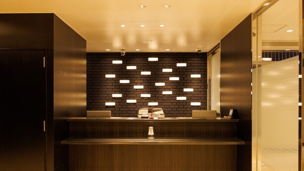 A reception area with a dark wood reception desk, a black brick wall with recessed lighting, and a tiled floor. - Floral Higashishinsaibashi Hotel (Osaka)