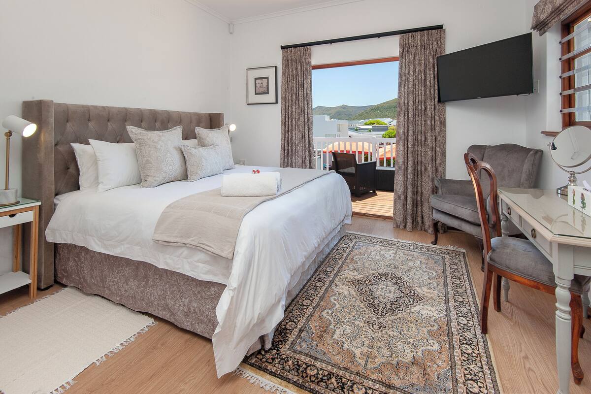 A bedroom with a large bed, a desk with a chair, a TV mounted on the wall, and a balcony with a view of the hills.