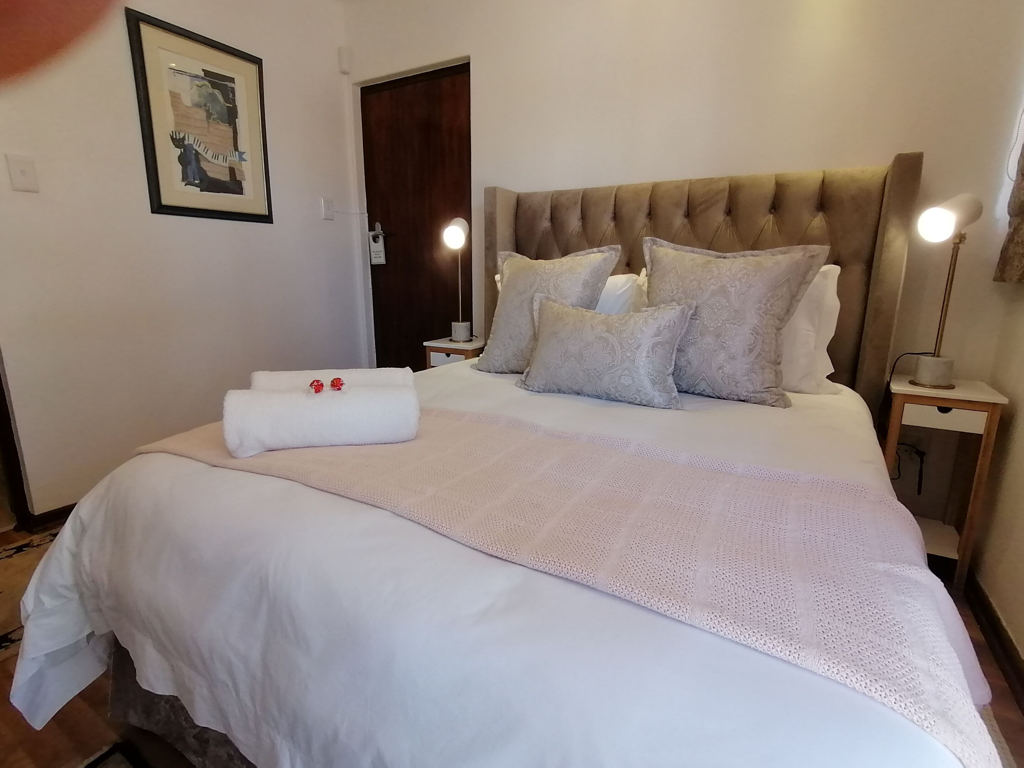 Una cama bien hecha con edredón blanco, varios almohadones y una toalla decorativa. Hay dos lámparas de noche y una mesita con una lámpara.