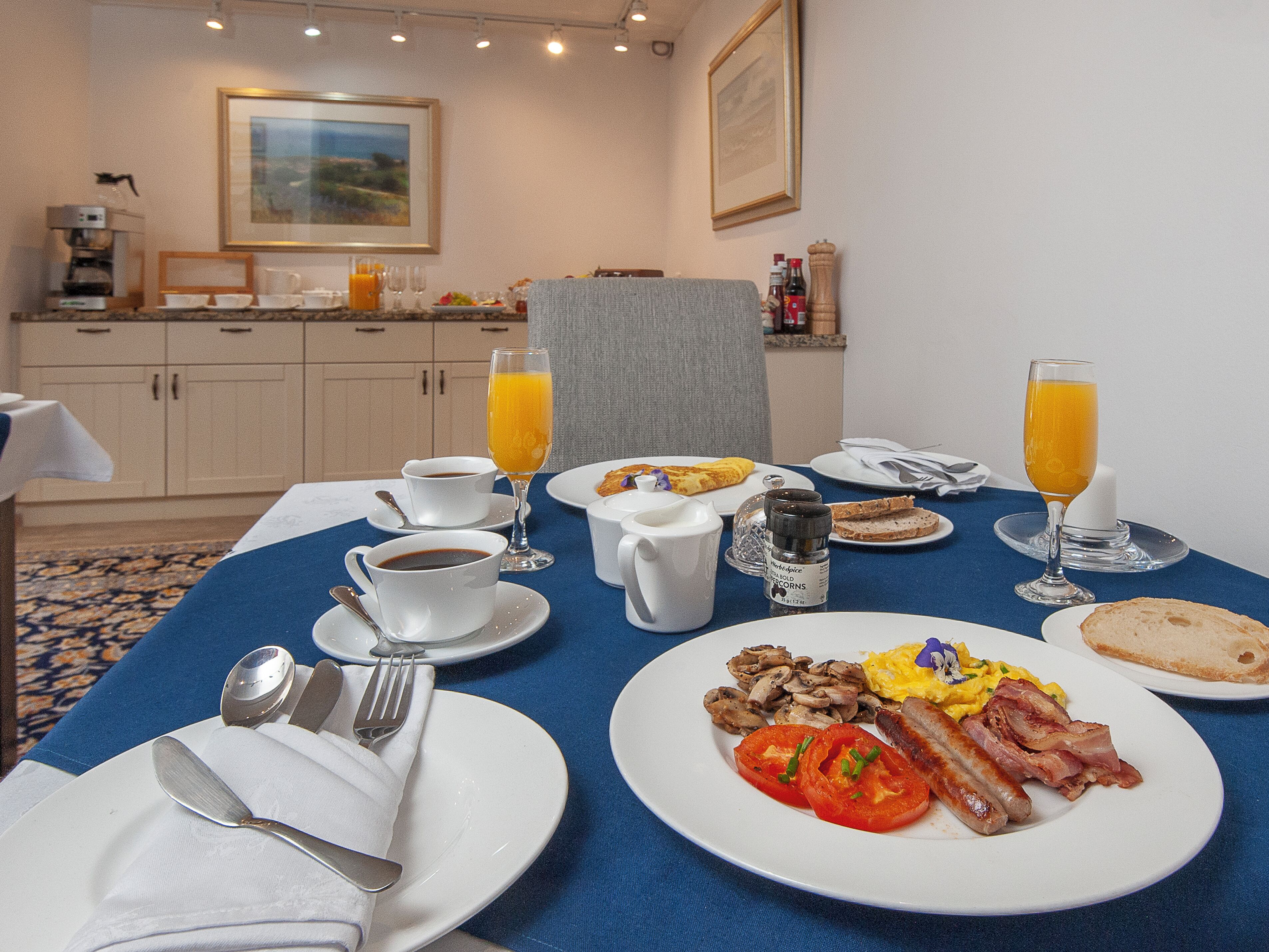 Una mesa de desayuno con platos de huevos, tocino, tomates y champiñones, junto con tazas de café y jugo de naranja.