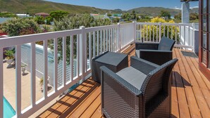 A wooden deck with wicker furniture and a white railing overlooking a pool and a residential area with hills in the background. - Villa Venusta (Hermanus)