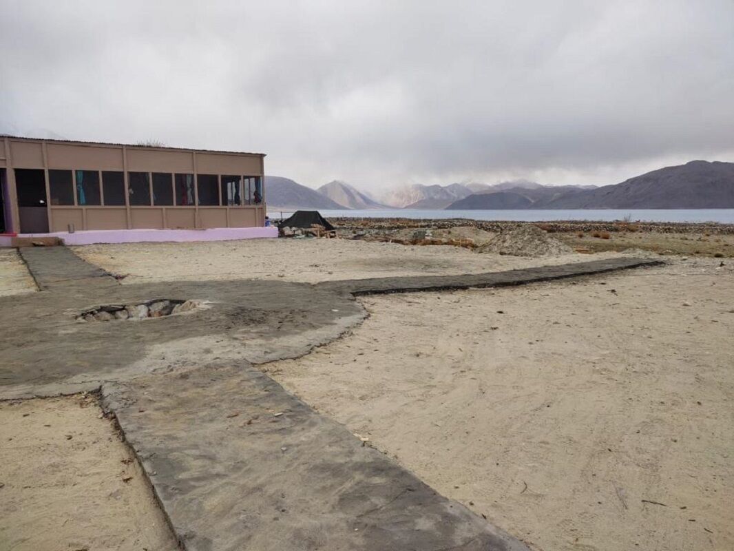 A building with a flat roof and large windows, situated on a sandy area with a concrete path leading to it.