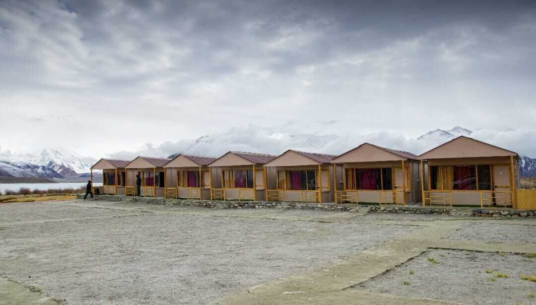 A row of simple, single-story buildings with large windows, set against a backdrop of mountains and a cloudy sky.