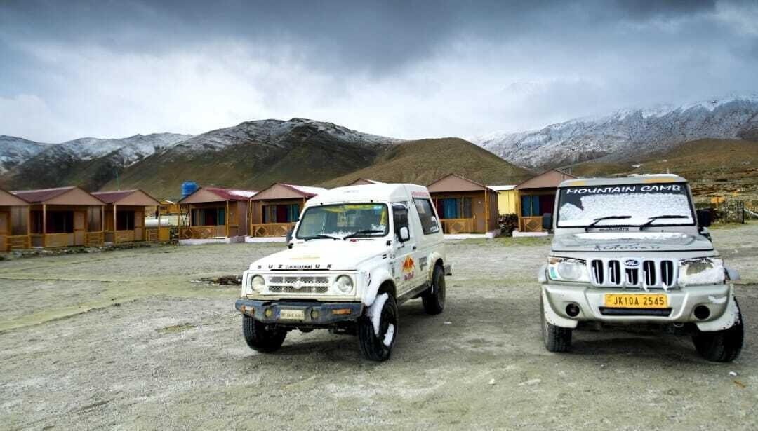 Deux SUV blancs sont stationnés devant une rangée de chalets en bois, avec une chaîne de montagnes en arrière-plan.