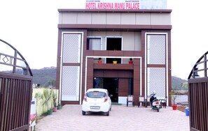 L’entrée de l’hôtel Krishna Manu Palace, avec une voiture blanche garée devant et une moto sur le côté droit.