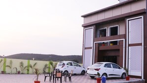 Uma área de estacionamento com dois carros brancos, plantas em vasos e um edifício com fachada estampada.