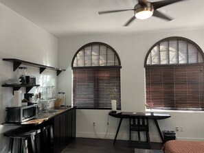 A compact kitchen with a microwave, toaster, and coffee maker. There are two windows with blinds, a ceiling fan, and a small dining table. - (Miami)