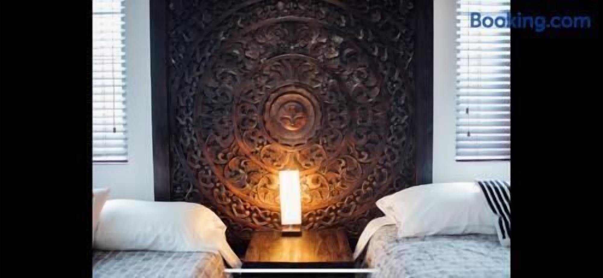 A room with a decorative headboard, a bedside table with a lamp, and two beds with pillows.