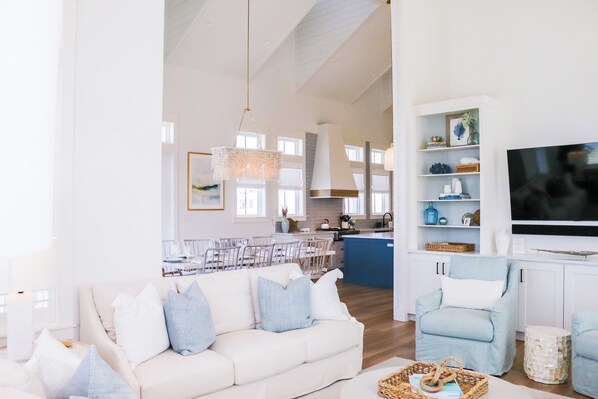 A spacious living room with a white sofa, a light blue armchair, and a television. A kitchen with white cabinets and a blue island is visible in the background. - Stunning Cinnamon Shore South Lakeside & Poolside Home With Amenities Galore (Port Aransas)