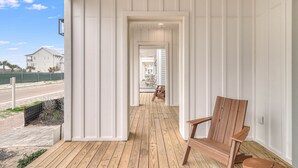 Una terraza de madera con una silla de madera, un muro exterior blanco y una vista de una casa al fondo.