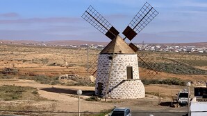Eine Windmühle mit steinernen Fundament und weißer Fassade, eingebettet in eine ländliche Landschaft unter blauem Himmel.
