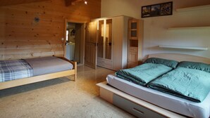 Ein Schlafzimmer einer Holzhütte mit zwei Betten, einem Kleiderschrank und einer durchgehenden Tür erkennbaren Küche.