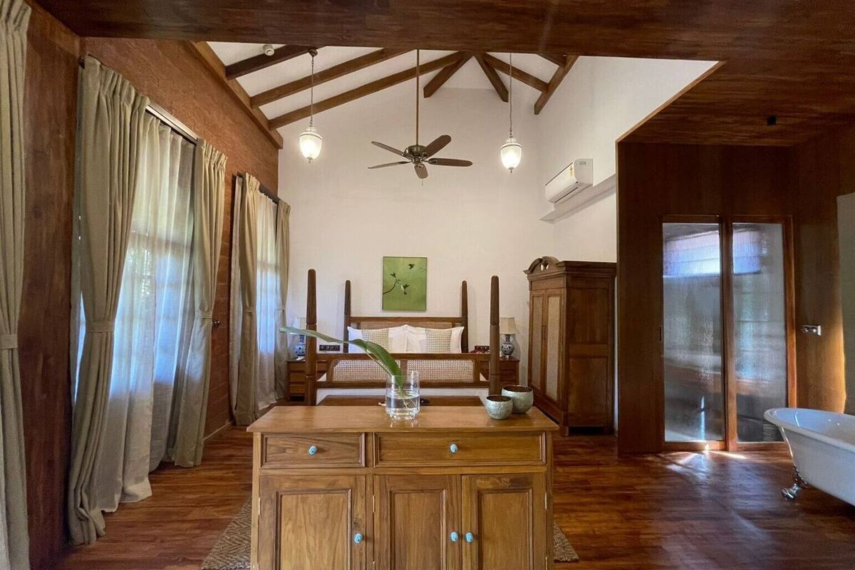 A bedroom with a wooden ceiling, a bed, a nightstand, a cabinet, a window with curtains, a ceiling fan, and a bathroom with a bathtub.