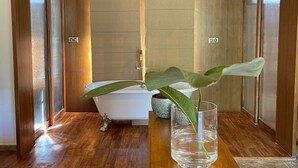 A modern bathroom with a wooden ceiling, a freestanding bathtub, and a glass vase with a plant on a wooden table. - The Postcard Saligao (Consua)