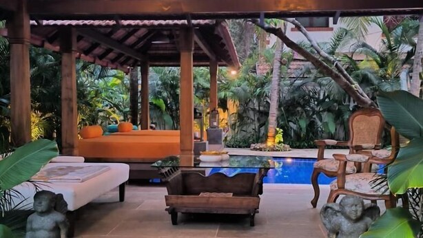 An outdoor lounge area with a pool, wooden furniture, and stone statues. - The Postcard Saligao (Consua)