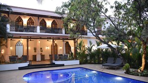 A pool area with a clear blue pool, lounge chairs, and a building with arched windows and a balcony. - The Postcard Saligao (Consua)
