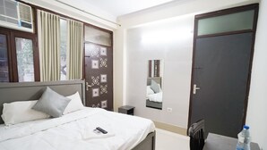 A neatly made bed with white bedding and pillows, a bedside table with a water bottle, a mirror on the wall, and a dark door. - Roomshala 034 Grihum Noida Sector 63 (Noida)
