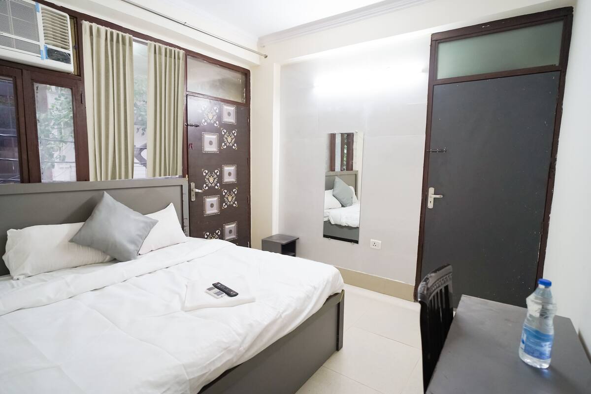 A neatly made bed with white bedding and pillows, a bedside table with a water bottle, a mirror on the wall, and a dark door.