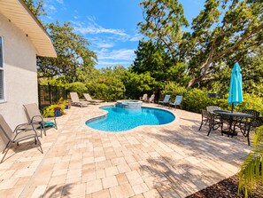 A swimming pool with a hot tub, surrounded by lounge chairs and a table with umbrellas, set against a backdrop of trees and a clear sky. - Paradise by the River - Relax in Style Near the Beach! Pool and Heated Spa (St. Augustine)