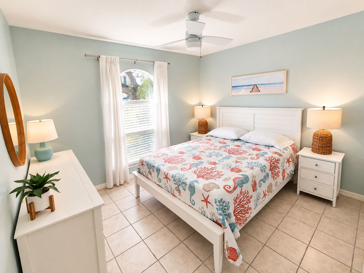 Une chambre à coucher comprenant un lit, des tables de chevet, un ventilateur de plafond, une fenêtre avec des rideaux, une plante et un miroir fixé au mur.