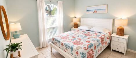 Une chambre à coucher comprenant un lit, des tables de chevet, un ventilateur de plafond, une fenêtre avec des rideaux, une plante et un miroir fixé au mur.